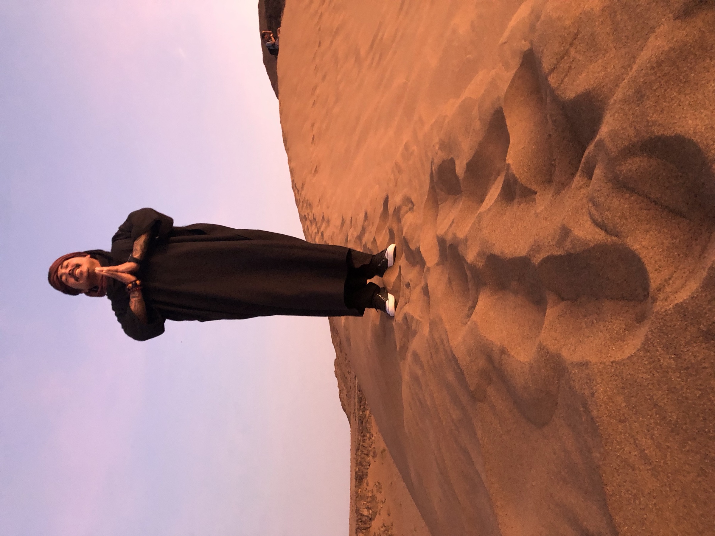 A lone figure on a sand dune, hands in prayer — the witness at distance.